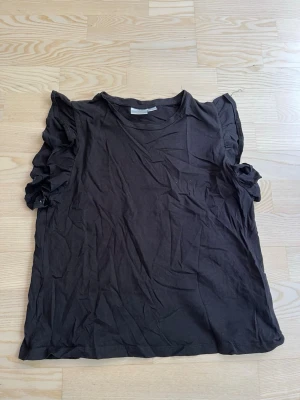 Svart topp med volangärmar - Snygg svart topp med volangdetaljer på ärmarna och rund halsringning. Perfekt för dig som vill ha en enkel men ändå trendig look. Tillverkad i mjuk bomull för skön känsla hela dagen.