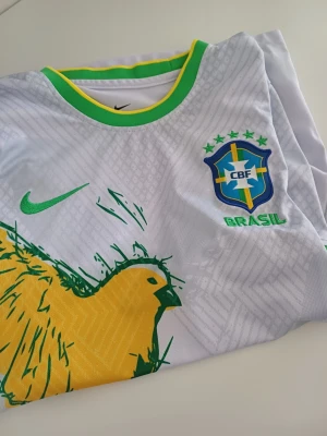 Vit Brasilien t-shirt Nike - Säljer en vit Brasilien t-shirt från Nike med gröna och gula detaljer. Tryck av en gul fågel på framsidan och broderad CBF-logga med texten BRASIL. Rund halsringning med grönt och gult kantband. Perfekt för dig som älskar fotboll och vill sticka ut.