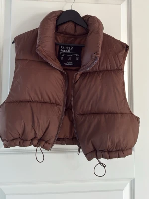 Brun dunväst med hög krage - Snygg brun dunväst med hög krage och dragkedja framtill. Västen har en puffig, quiltad design och justerbar dragsko i nederkant för perfekt passform. Perfekt för lager-på-lager och ger en trendig look till din outfit.