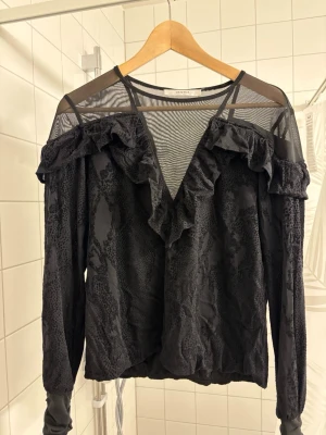 Svart blus med volanger och meshdetaljer - Snygg svart blus med transparent mesh upptill och volangdetaljer längs axlar och V-ringning. Långärmad modell med lätt mönstrad struktur i tyget. Perfekt för dig som vill ha en trendig och unik look.