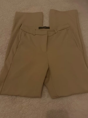 Beige kostymbyxor från Vero Moda - Snygga beige kostymbyxor från Vero Moda med raka ben och pressveck. Byxorna har hällor för skärp och sidofickor. Perfekta för en clean och stilren look. Materialet känns mjukt och har en lätt stretch.