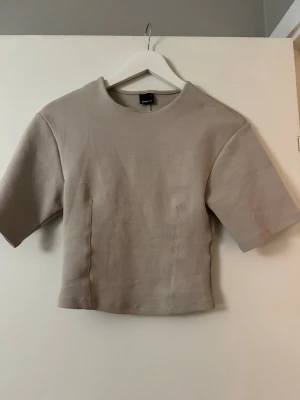 Beige topp med kort ärm - Säljer en stilren beige croppad topp med korta ärmar och rund hals. Toppen har en figursydd passform och är gjord i ett mjukt material som känns skönt mot huden. Perfekt att matcha med höga jeans eller kjol för en clean look.
