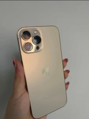Guldfärgad iPhone med trippelkamera - Snygg guldfärgad iPhone med stilren design och platt baksida i glas. Telefonen har tre stora kameralinser i hörnet och metallram runt om. Apple-loggan syns tydligt på baksidan. Perfekt för dig som gillar modern teknik och elegant look. 77% batterihälsa. Inga sprickor eller repor. Funkar perfekt 