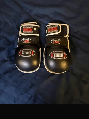 Svart kampsportsutrustning Fighter - Säljer ett set svart kampsportsutrustning från Fighter, inklusive ett par mma och benskydd. Handskarna har kardborreband och vit kant, medan benskydden har flera justerbara remmar och tydlig logga. Perfekt för träning inom kampsport. Aldrig använda nypris 1600