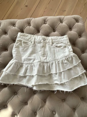 Vit volangkjol med jeanslook - Supersöt vit kortkjol i jeansmaterial med två volanglager och råa kanter. Kjolen har klassiska fem fickor och bälteshällor i midjan. Perfekt för dig som vill ha en trendig och avslappnad stil.