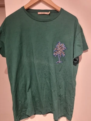 Grön t-shirt från Whyred - Snygg grön t-shirt med ett coolt palmtryck i blått och vitt på bröstet. Klassisk rund halsringning och korta ärmar. Tillverkad i mjuk bomull som känns skön mot huden. Perfekt för dig som gillar avslappnad stil med en twist.