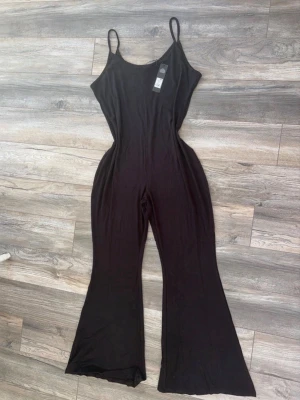 Svart jumpsuit från Fashion Nova - Snygg svart jumpsuit från Fashion Nova med smala axelband och utsvängda ben. Tillverkad i mjukt stretchigt tyg som sitter skönt på kroppen. Perfekt för dig som gillar en enkel men trendig look. Enkel att styla med sneakers eller klackar.