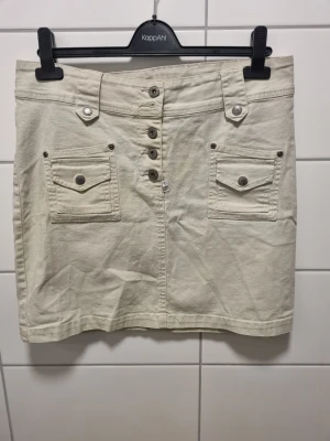 Beige kortkjol med knappar från Lindex - Stilren beige kortkjol från Lindex med framfickor och dekorativa knappar framtill. Kjolen har bälteshällor och är tillverkad i ett mjukt bomullsmaterial. Perfekt för dig som gillar en enkel men snygg look.