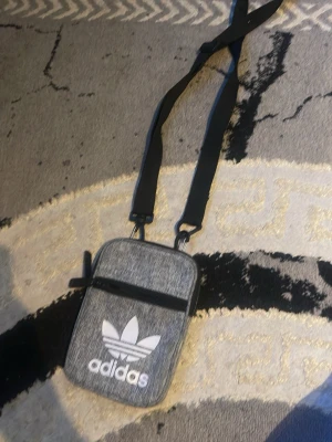 Grå axelväska från Adidas - Snygg grå axelväska från Adidas med klassiska vita loggan framtill. Väskan har justerbar svart axelrem och en praktisk dragkedjeficka på framsidan. Perfekt för att bära det viktigaste när du är på språng.