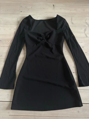 Svart långärmad kort klänning - Stilren svart klänning med lång ärm och twistad detalj vid bysten. Klänningen har en figurnära passform och är kort i modellen, perfekt för en trendig look. Tillverkad i ett mjukt och stretchigt material som ger en bekväm känsla.