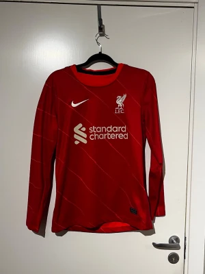 Liverpool långärmad matchtröja Nike - Röd långärmad Liverpool FC matchtröja från Nike med vita detaljer och klubbmärke på bröstet. Tröjan har tryckt sponsorlogga och tunna diagonala linjer över hela plagget. Perfekt för dig som älskar fotboll och vill bära ditt lag med stil.