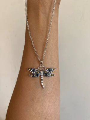 Halsband med trollslända i silverfärg - Unikt halsband med en trollslända som hänge. Hänget är detaljerat med svarta, vita och gula inslag på vingarna och har en silverfärgad kedja. Perfekt för dig som gillar naturinspirerade smycken och vill sticka ut med en cool accessoar.
