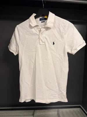 Vit pikétröja från Polo Ralph Lauren - Klassisk vit pikétröja från Polo Ralph Lauren i slim fit-modell. Tröjan har korta ärmar, krage och knappar framtill samt den ikoniska broderade loggan på bröstet. Tillverkad i mjuk bomull som känns skön mot huden.
