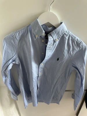 Ljusblå skjorta från Ralph Lauren - Klassisk ljusblå skjorta från Ralph Lauren med button-down krage och broderad logga på bröstet. Skjortan har långa ärmar och knappar framtill. Perfekt till jeans eller chinos för en clean och stilren look.