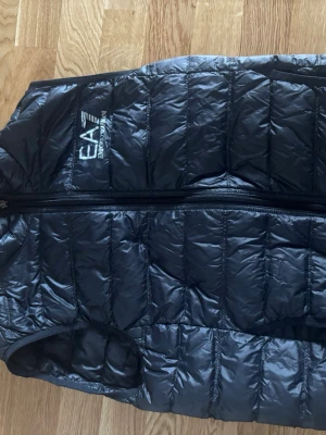 Svart dunväst från EA7 Emporio Armani - Snygg svart dunväst från EA7 Emporio Armani med quiltad design och dragkedja framtill. Västen har hög krage och logga på bröstet. Perfekt för lager på lager och ger en sportig look.