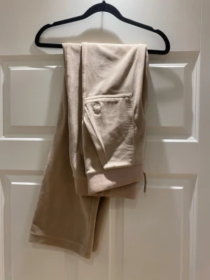 Beige mjukisbyxor med fickor - Snygga beige mjukisbyxor med resår i midjan och muddar vid bensluten. Byxorna har en praktisk ficka på sidan och är gjorda i ett mjukt och skönt material som passar perfekt för en avslappnad stil.