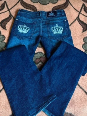 Blå bootcut jeans med kronbrodyr - Säljer ett par blå bootcut jeans med snygga broderade kronor på bakfickorna. Jeansen har klassisk femficksmodell och är tillverkade i denim med lätt stretch. Perfekta för dig som gillar detaljer och vill sticka ut lite extra.