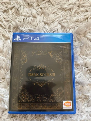Dark Souls III till PS4 - Dark Souls III till PlayStation 4, komplett med fodral och skiva. Omslaget är svart med guldiga detaljer och spelets logga. Skivan har en mörk design med spelets titel och åldersmärkning. Perfekt för dig som gillar utmanande action-RPG.