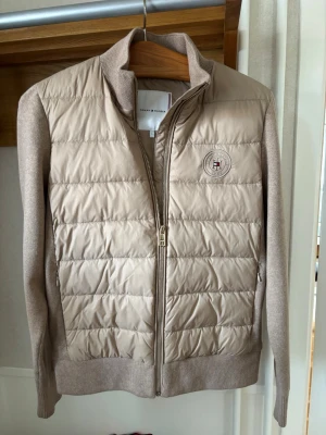 Cardiganjacka Tommy Hilfiger - Hej säljer min snygga beige cardigan jacka från Tommy Hilfiger med dragkedja framtill, hög krage och ribbade muddar. Framsidan är vadderad medan ärmar och rygg är stickade. Märkeslogga på bröstet för extra detalj. Är i mycket bra skick utan defekter. Priset för att diskutera. Hör av er vid frågor!