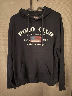 Marinblå hoodie Polo   - Tja! Jag säljer en marinblå hoodie från Polo Club med vit text och amerikansk flagga på bröstet. Klassisk känguruficka framtill och justerbar huva med snören. Perfekt för en avslappnad och sportig stil, bara att hojta till vid funderingar:)