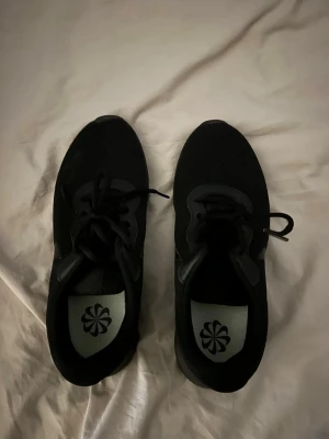 Svarta Nike sneakers med snörning - Säljer ett par helsvarta Nike sneakers med klassisk snörning och diskret swoosh på sidan. Skorna har en mesh-liknande ovandel och en bekväm, tjock sula som passar perfekt till streetwear och vardagsstil.