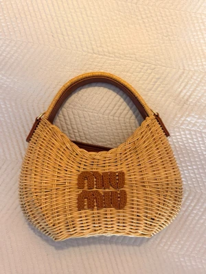 Flätad handväska från Miu Miu - Snygg flätad handväska från Miu Miu i naturfärgad rotting med bruna detaljer och handtag. Väskan har en rundad form och logotypen är broderad framtill. Perfekt accessoar för en trendig och avslappnad look.