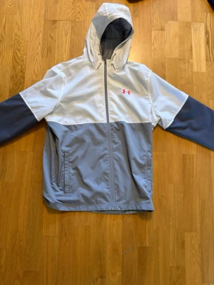 Under Armour tvåfärgad vindjacka - Snygg vindjacka från Under Armour i ljusgrått och vitt med rosa logga på bröstet. Jackan har huva, dragkedja framtill och två sidofickor. Perfekt för blåsiga dagar och sportiga outfits.