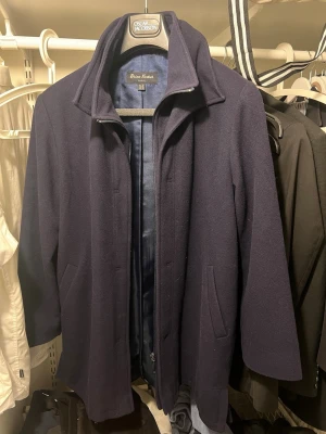 Jacka/Kappa - Stilren mörkblå ullkappa från Brian Tucker  med klassisk krage och diskreta sidofickor. Kappan har rak passform och helfodrad insida, perfekt för kyliga dagar. Enkel design som funkar till många olika looks.