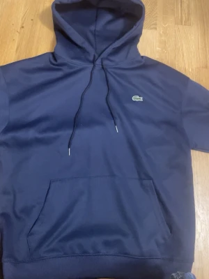 Mörkblå hoodie från Lacoste - Snygg mörkblå hoodie från Lacoste med klassisk krokodillogo på bröstet. Hoodien har huva med dragsko och en stor magficka framtill. Tillverkad i mjukt material som känns skönt mot huden. Perfekt för en avslappnad och stilren look.