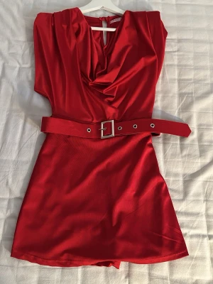 Röd kort jumpsuit med bälte - Snygg röd jumpsuit i glansigt tyg med draperad urringning och matchande bälte i midjan. Kort modell med vida ben och ärmlös design. Perfekt för dig som vill sticka ut med en färgstark och trendig look. passar 36-38