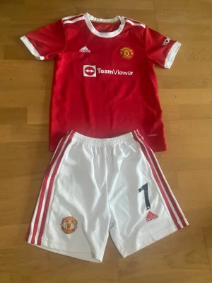 Manchester United Ronaldo matchställ - Säljer ett Manchester United matchställ med Ronaldo 7. Tröjan är röd med vita detaljer och klubbmärke, medan shortsen är vita med röda ränder och nummer 7. Båda delarna har Adidas-logga och är tillverkade i lätt, andningsbart material.