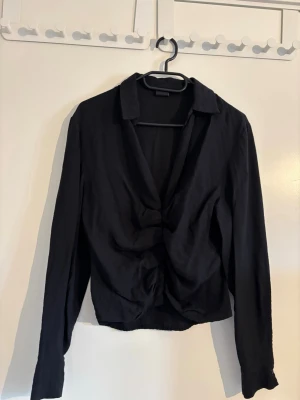 Svart blus med twistad front - Snygg svart blus med twistad detalj framtill och djup v-ringning. Blusen har krage och långa ärmar, vilket ger en stilren och trendig look. Tillverkad i ett mjukt, tunt tyg som faller fint. Perfekt att matcha med jeans eller kjol.