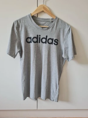 Grå Adidas t-shirt med logga strl S - Snygg grå t-shirt från Adidas med stor svart logga på bröstet. Klassisk rund halsringning och korta ärmar. Material 87% bomull, 13% polyester. Storlek S.