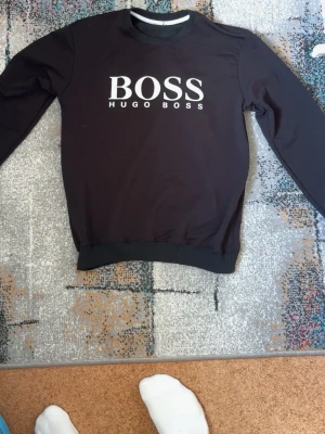 Svart sweatshirt från Hugo Boss - Stilren svart sweatshirt från Hugo Boss med stor vit logga på bröstet. Tröjan har rund halsringning, långa ärmar och ribbade muddar vid ärmslut och nederkant. Perfekt för en avslappnad och trendig look.