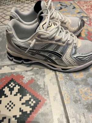 Asics gel kayano 14 white/midnight - Snygga sneakers i vitt och silver med svarta detaljer och meshpaneler för extra ventilation. Skorna har en sportig design med tydliga linjer och tjock sula som ger bra stöd. Perfekta för dig som gillar en modern och fräsch look.