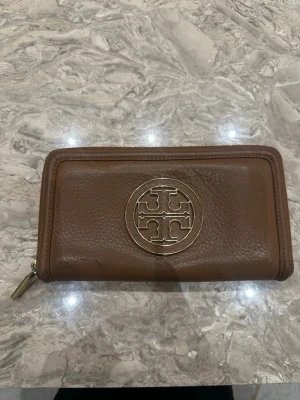 Brun plånbok med gulddetalj från tory burch - Snygg brun plånbok i läder med en stor guldfärgad metallogo på framsidan. Plånboken har dragkedja runtom och en stilren, rektangulär form. Perfekt för dig som gillar klassisk och elegant design.