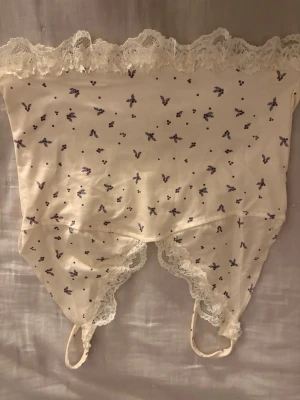 Vit spetsbody med små lila blommor - Söt vit body med tunna axelband och spetsdetaljer längs kanterna. Tyg med små lila blommor och prickar ger en romantisk känsla. Perfekt för dig som gillar feminina och mjuka underkläder. Materialet känns lent och stretchigt.