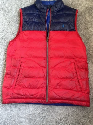 Röd och marinblå dunväst - Snygg dunväst i rött med marinblå axelparti och ståkrage. Västen har två framfickor med dragkedja och quiltad design. Perfekt för lager-på-lager och ger en sportig look. Materialet är glansigt och västen stängs med dragkedja framtill. Den passar dig mellan 155-165. Den har tyvärr ett litet hål inuti västen men syns knappt 