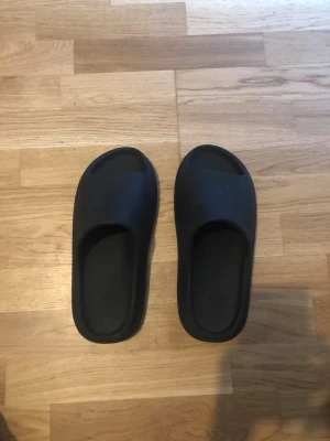 Svarta Yeezy Slides - Säljer ett par svarta yeezy slides med chunky sula och öppen tå. Tillverkade i mjukt gummi för extra komfort och har en modern, minimalistisk design. Perfekta för att glida runt hemma eller på stranden.