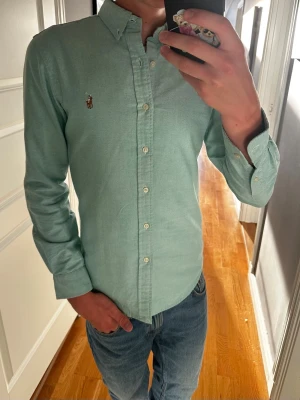Grön Oxford Ralph Lauren skjorta - Klassisk och stilren skjorta från Polo Ralph Lauren som passar både till vardags och lite mer uppklädda outfits. Jag är 180 cm / 70 kg och bär storlek S på bilderna.  Skjortan är i mycket fint skick utan defekter. Inga fläckar, hål eller andra skador.  Modell: Polo Ralph Lauren Oxford Shirt Storlek: S Nypris: ca 1 550 kr Skick: Mycket fint skick  Mått:  * Längd: 70 cm * Bredd: 45 cm * Ärmlängd: 61 cm  📦 Postas inom 24 timmar! 📸 Se bilder för skick och passform.