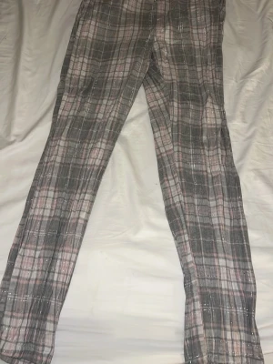 Rutiga pyjamasbyxor från KappAhl - Mysiga pyjamasbyxor från KappAhl i grått och rosa rutigt mönster med tunna silvertrådar. Byxorna har en söt rosett framtill i midjan och resår för extra komfort. Perfekta för chillkvällar hemma.