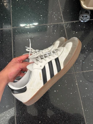 Adidas Samba vita och svarta sneakers - Klassiska Adidas Samba sneakers i vitt och svart med bruna gummisulor. Skorna har tre svarta ränder på sidan, grå mockadetaljer vid tån och vita skosnören. Perfekta för en sportig och retro look.