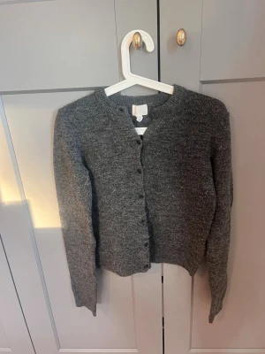 Grå kofta från H&M - Mysig gråmelerad kofta från H&M i storlek XS. Koftan har rund halsringning, knäppning framtill med matchande knappar och långa ärmar. Perfekt att slänga över en t-shirt eller linne för en enkel och stilren look.