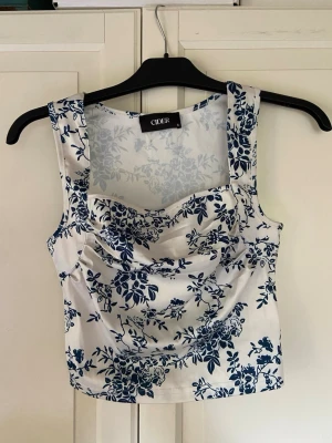 Vit blommig linnetopp från Cider - Supersnygg vit linnetopp från Cider med blått blommigt mönster. Toppen har breda axelband, draperad framsida och är ärmlös. Perfekt för sommarens alla outfits och ger en fräsch och trendig look.