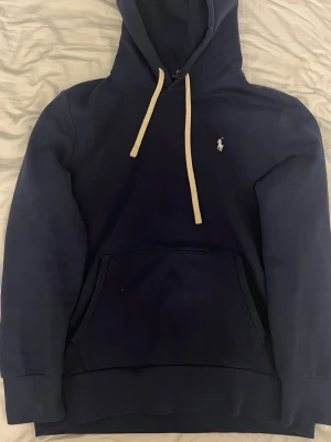 Polo Ralph Lauren hoodie - Mörkblå hoodie med vit snörning och klassisk känguruficka framtill. Liten broderad logga på bröstet. Mjuk och skön, perfekt för chill dagar. Huva och långa ärmar ger en avslappnad vibe.