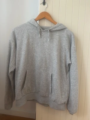 Grå basic hoodie med huva - En enkel och stilren grå hoodie med huva och dragsnören. Hoodien har en klassisk känguruficka framtill, ribbade muddar vid ärmslut och nederkant. Perfekt för en avslappnad och bekväm look. Materialet känns mjukt och skönt mot huden.