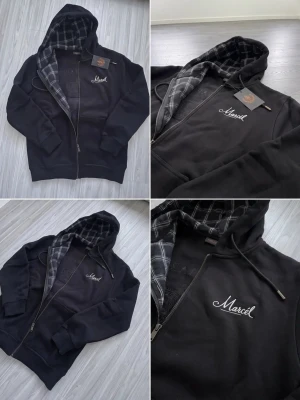 Svart hoodie med rutigt foder och dragkedja - Snygg svart hoodie med dragkedja och huva som har rutigt foder i grått och svart. På bröstet finns broderad text 'Marcel' i vitt. Hoodien har fickor framtill och justerbara snören i huvan. Perfekt för en avslappnad streetwear-look.