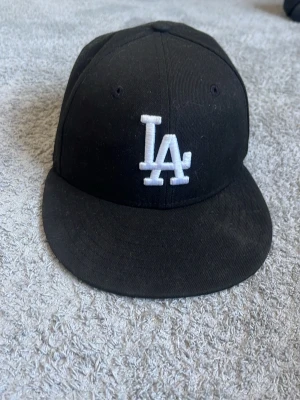Svart LA-keps från New Era - Säljer en svart keps från New Era med LA-logga broderad i vitt framtill. Klassisk 59FIFTY-modell med rak skärm och strukturerad passform. Tillverkad i bomullsmix och perfekt för dig som gillar streetwear och sportig stil.
