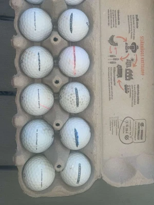 Vita golfbollar i äggkartong - Säljer ett set med vita golfbollar, flera av märket super Soft. Bollarna har klassisk rund form och vissa har färgade markeringar. Perfekt för golfträning eller spel på banan.
