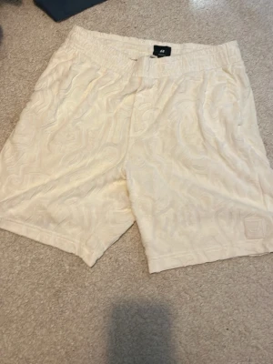 Beige mönstrade shorts från H&M - Säljer ett par beige shorts från H&M med subtilt mönster i tyget. De har elastisk midja och fickor både fram och bak. Perfekta för varma dagar och har en avslappnad passform.